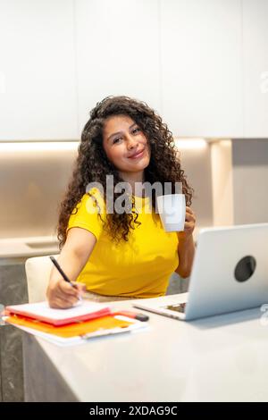 Vertical photo of a female latin content creator freelancer writing notes and drinking coffee working from home Stock Photo