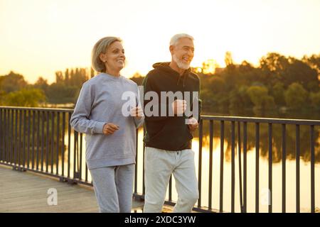 Sporty senior smiling couple in sportswear jogging along a bridge in park having sport workout. Stock Photo