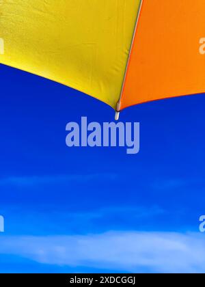 A close-up shot of a bright yellow and orange beach umbrella against a clear blue sky with wispy white clouds. The umbrella is positioned so that only Stock Photo