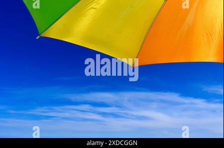 A close-up shot of a bright yellow and orange beach umbrella against a clear blue sky with wispy white clouds. The umbrella is positioned so that only Stock Photo