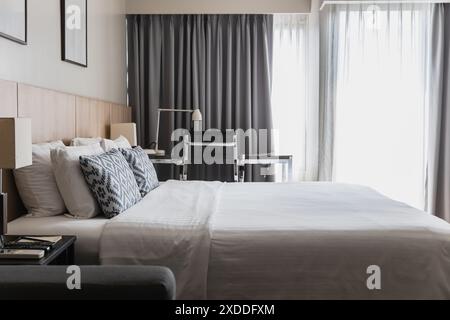 Modern bedroom featuring a neat bed, decorative pillows, a sleek desk, and large windows with curtains. Ideal for concepts of comfort, style, and cont Stock Photo