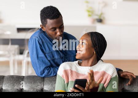 Sitting on couch, woman holding smartphone and smiling at man leaning over backrest Stock Photo