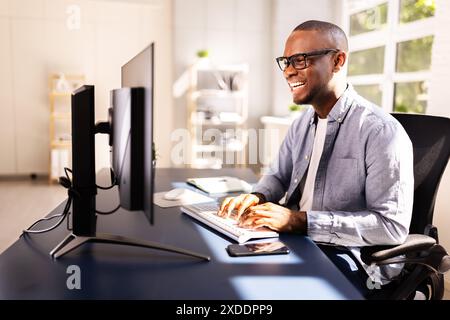 Efficient African American Businessman Managing Documents and Multiple Computer Screens at Corporate Office Desk, Smiling While Working. Stock Photo