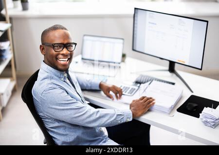 Top African-American Tax Advisor Smiling While Multitasking at His Desk in a Corporate Office. Professional Accountant Analyzing Financial Data. Stock Photo