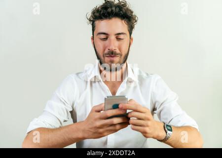 A young man in a white shirt using a smartphone, looking focused and engaged, indoor setting, modern communication, technology usage, casual attire, d Stock Photo