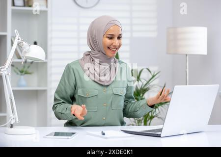 Smiling young muslim woman in hijab working in office, sitting at desk and talking on video call via laptop. Stock Photo