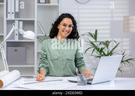 Portrait of a young Muslim woman architect and engineer working in the office with a laptop and with blueprints, sitting at a table with a laptop, looking and smiling at the camera. Stock Photo