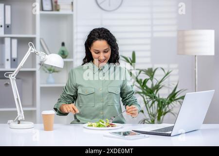 Lunch break at work. Young Latina woman sitting at desk on laptop in office, drinking coffee and eating salad. Stock Photo