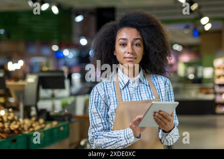 Portrait of young smiling African American woman, supermarket owner and worker standing in grocery department in apron, holding and using tablet, looking at camera. Stock Photo