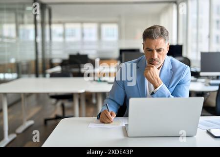 Busy serious middle aged business man working on laptop in office writing notes. Stock Photo
