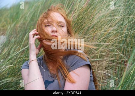 young red-haired woman with the wind blowing her hair into her face sits on vacation in the windy sand dunes of St. Peter Ording, copy space Stock Photo