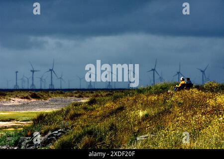 Couple sitting looking out to sea on Haverigg Beach near Millom UK with a large offshore wind farm on the horizon. Stock Photo