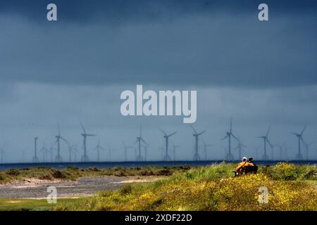 Couple sitting looking out to sea on Haverigg Beach near Millom UK with a large offshore wind farm on the horizon. Stock Photo