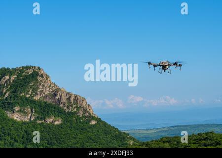 An agricultural drone flies over a mountainous landscape on a clear, sunny day. The drone is in the foreground, with the mountains and forested hills in the background. Stock Photo