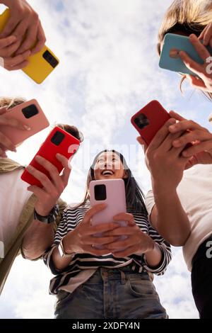 Low angle view of group happy unrecognizable people in circle using colorful mobiles phones outdoor. Stock Photo