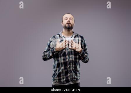 Bearded man with closed eyes holding both hands on his chest, experiencing discomfort and pain. He is standing on a gray background Stock Photo