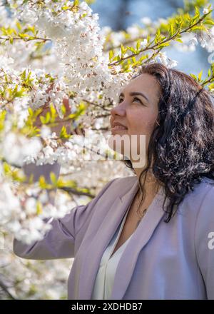 romantic image of a stylish woman in a light jacket. Positive, spring mood. A cute girl gently holds a white sakura branch and looks at the flowers, s Stock Photo