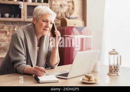 Senior Woman Talking on Phone, Writing Notes While Using Laptop Stock Photo