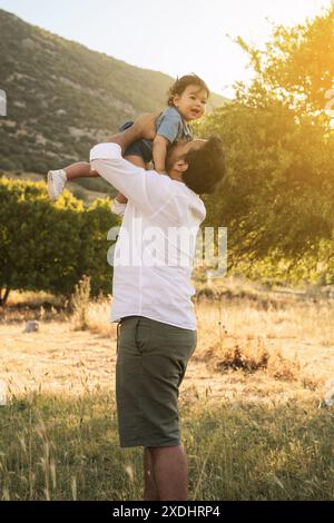 A father in a white shirt playfully lifting his one-year-old son in the air in a grassy natural setting. A tree and a grassy field form the backdrop Stock Photo