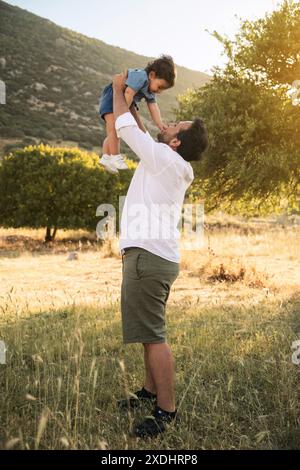 A father in a white shirt playfully lifting his one-year-old son in the air in a grassy natural setting. A tree and a grassy field form the backdrop Stock Photo