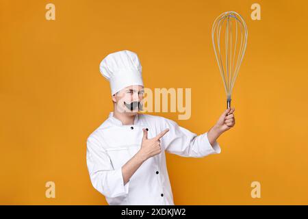 Pastry chef with fake mustache pointing at big whisk on orange background Stock Photo