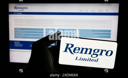 Person holding mobile phone with logo of South African investment company Remgro Limited in front of business web page. Focus on phone display. Stock Photo