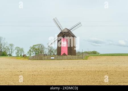A solitary traditional windmill with red details stands in a plowed field, under a cloudy sky. Stock Photo