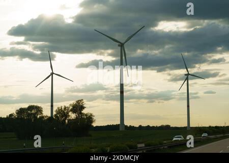 A car travels down a road lined with towering wind turbines. The vehicle moves alongside the rotating blades generating renewable energy. Stock Photo