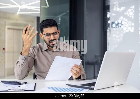 Young man wearing glasses and looking stressed and frustrated while holding paperwork in an office setting. He is seated at a desk with a laptop, documents. Emotion of worry and confusion. Stock Photo