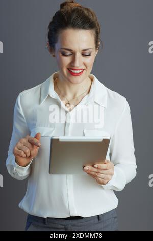 happy trendy female employee in white blouse using tablet PC against grey background. Stock Photo