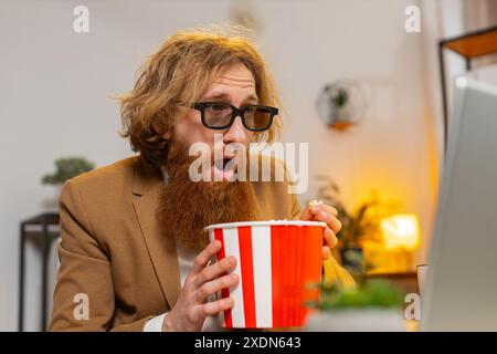 Caucasian businessman freelancer taking break from work wearing 3D glasses earing popcorn and watching movie on laptop. Smiling ginger man guy sitting at home office desk having snacks during break. Stock Photo