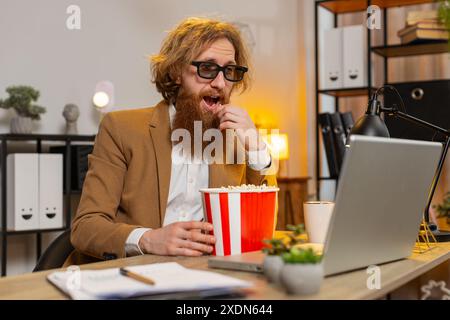 Caucasian businessman freelancer taking break from work wearing 3D glasses earing popcorn and watching movie on laptop. Smiling redhead man guy sitting at home office desk having snacks during break. Stock Photo