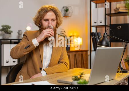 Focused Caucasian businessman sitting at workplace desk drinking water while working with laptop in home office. Millennial redhead employee freelancer looks at computer screen browse surf internet. Stock Photo
