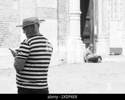 Venice, Italy - June 30th 20220 Gondolier wearing a striped shirt and a straw hat using a smartphone in venice, italy, with a beggar sitting in the ba Stock Photo