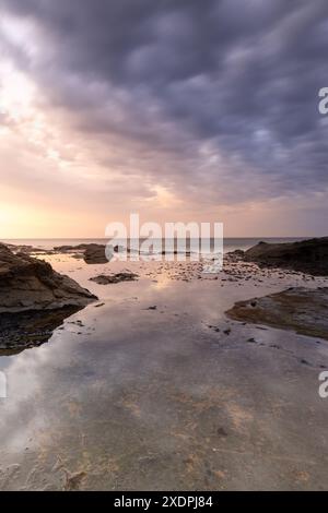 Rocky shoreline under a dramatic sunset sky reflecting on wet sand Stock Photo