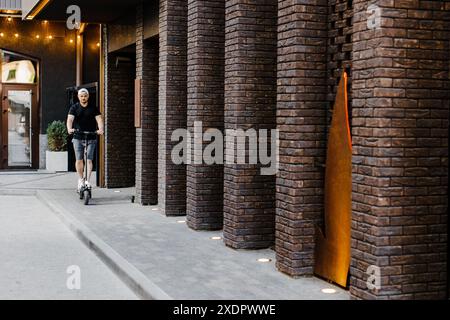 Young handsome man in white baseball cap riding on scooter to work along the street in the city. Active lifestyle, backpacker traveler. Ecology and cl Stock Photo