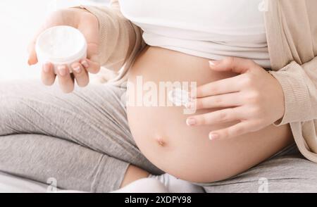 Pregnant Woman Applying Moisturizer Cream on Belly in Cozy Bedroom Stock Photo