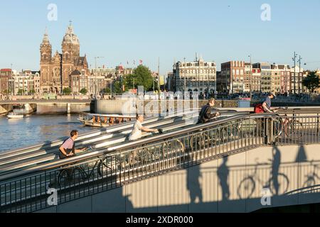 Cyclists push their bicycles up the travelator from the underground bicycle parking facility at Centraal Station, with the canal and the basilica of Saint Nicholas in the background, in Amsterdam, Netherlands. Stock Photo