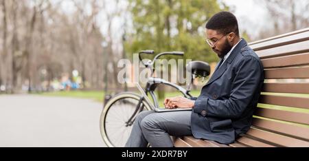 Businessman using laptop on park bench near bicycle Stock Photo