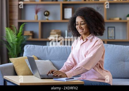 Smiling woman working from home on laptop seated on sofa in cozy living room. Concept of remote work, home office, comfort Stock Photo