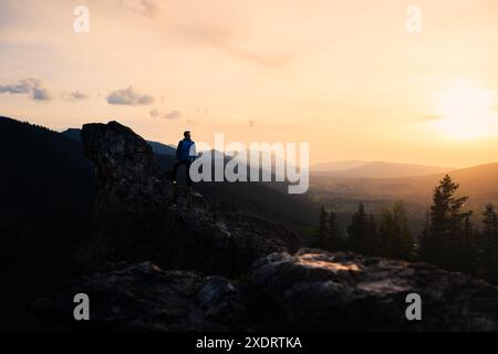 At dusk, a person stands on a mountain peak, surrounded by the natural landscape. The sky is painted with vibrant colors of the sunset Stock Photo