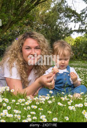 Portrait of mother and little son. A happy mother with wavy hair lies on the green grass among white daisies, a small baby with wavy, blond hair sits Stock Photo