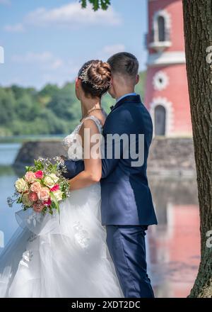 Young beautiful bride and groom stand in front of the water and look at the pink lighthouse. Rear view Newlyweds on a walk. Wedding, love, married cou Stock Photo