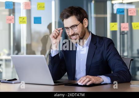 Smiling businessman working on laptop in modern office with sticky notes on glass wall. Professional in business attire focusing on work, planning, and strategy. Bright, collaborative workspace Stock Photo