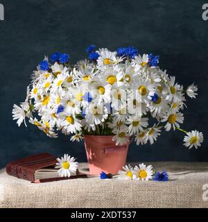 Summer still life. A large bouquet of daisies and cornflowers in a pink bucket. Nearby is a notebook and a pencil on a blue background. Stock Photo