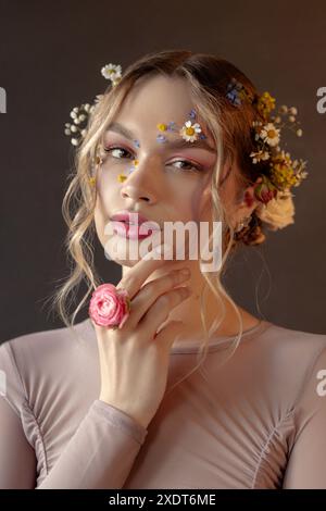 A woman with blonde hair and flowers in her hair and on her face, wearing a light pink long-sleeved shirt, looks directly at the camera. Stock Photo