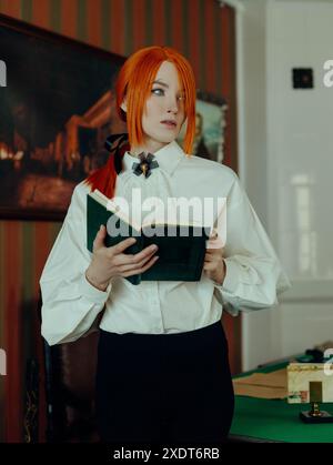 Woman with long, bright red hair and green eyes is standing in a vintage interior, wearing a white button-down shirt and black trousers, holding an op Stock Photo