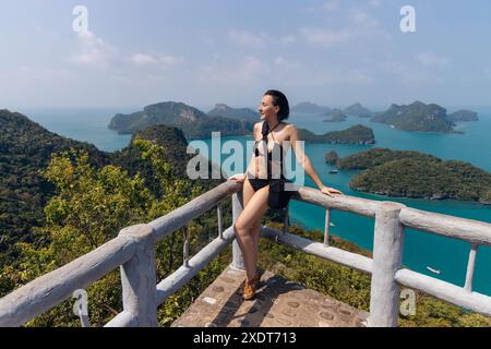 Woman in a black bikini stands on a wooden railing overlooking a stunning panorama of emerald green water and lush islands in Thailand. Stock Photo