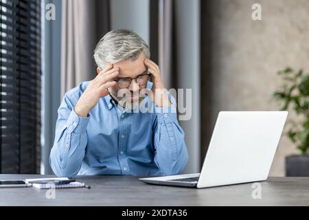 Stressed businessman wearing glasses working on laptop in office. Man feeling overwhelmed by work, holding head in hands. Modern office setting with natural light Stock Photo