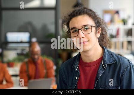 Candid close up portrait of long haired young man wearing glasses looking at camera in office and smiling internship concept copy space Stock Photo